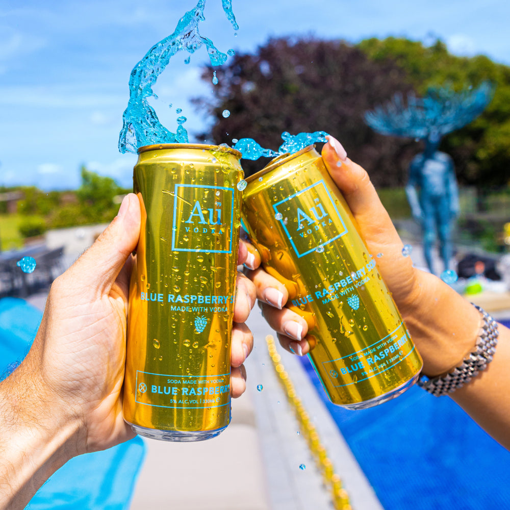 Two hands holding Au Vodka Blue Raspberry vodka cocktail cans with blue liquid splashing outdoors