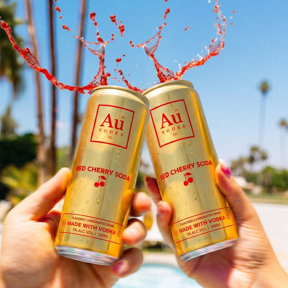 Two gold Au Vodka Red Cherry Soda cans with red liquid splashing from the tops